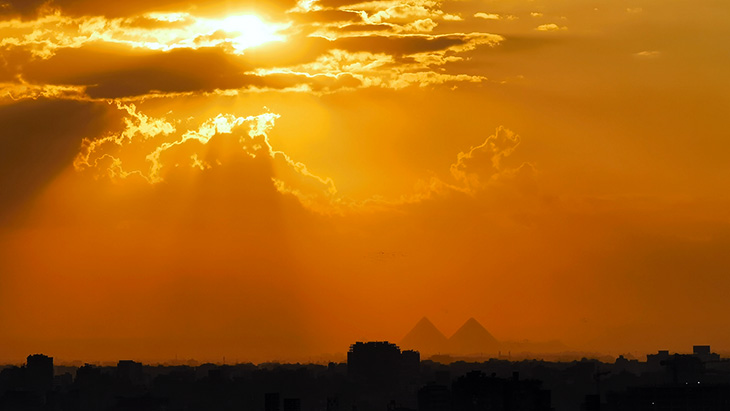 Vue sur Le Caire avec les pyramides du plateau de Guizeh en fond. Le soleil est brûlant au-dessus des nuages et donne une atmosphère rougeoyante au paysage avec la ville dans la pénombre.